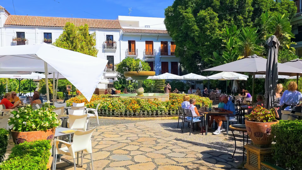 Café terraces surrounding a fountain and flowerbeds in an Estepona old town square.