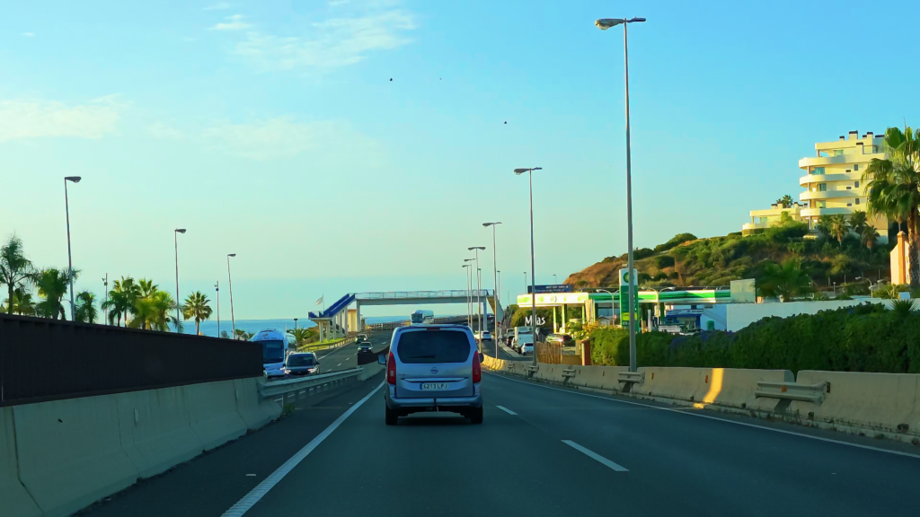 Cars travelling along a coastal road towards Estepona, with buildings and palm trees alongside.